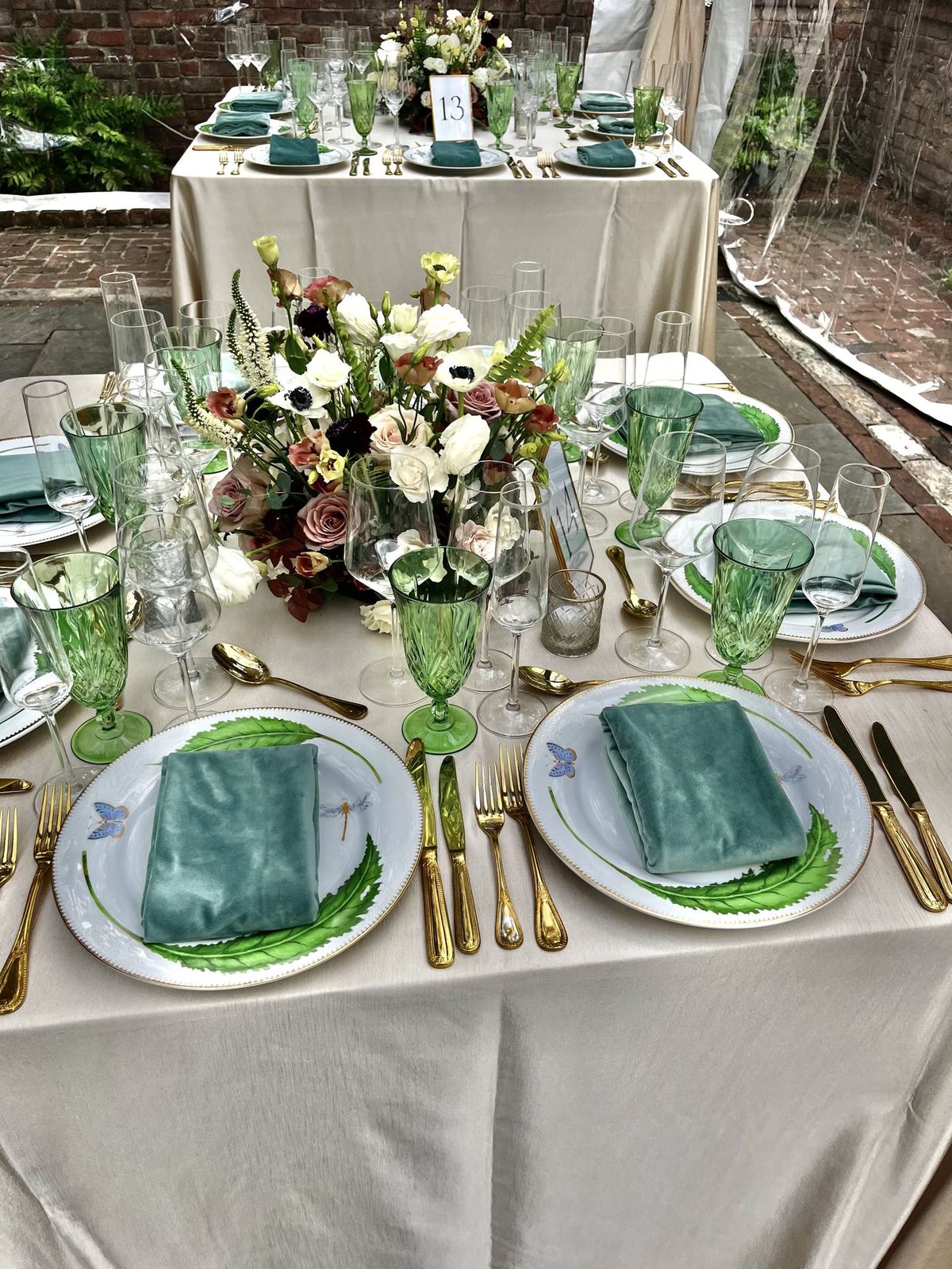 Courtyard tablescape with green goblets, velvet napkins, gold flatware, and a garden rose centerpiece